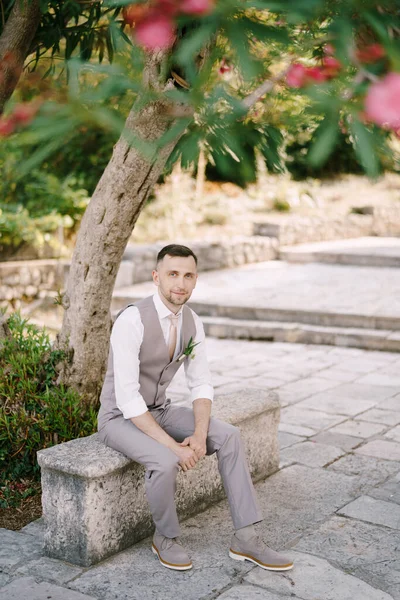 Brudgummen sitter på en stenbård under ett blommande träd i trädgården — Stockfoto