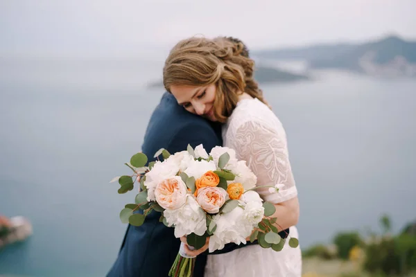 Pengantin pria memeluk pengantin wanita dengan karangan bunga sambil berdiri di gunung di atas laut. Potret — Stok Foto