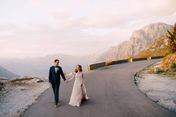 Bruid en bruidegom lopen langs de asfaltweg tegen de achtergrond van bergen — Stockfoto