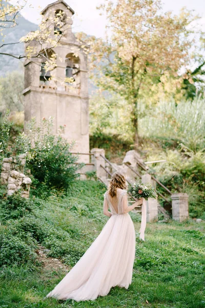 Novia con un ramo camina a lo largo de la hierba verde a la antigua capilla. Vista trasera —  Fotos de Stock