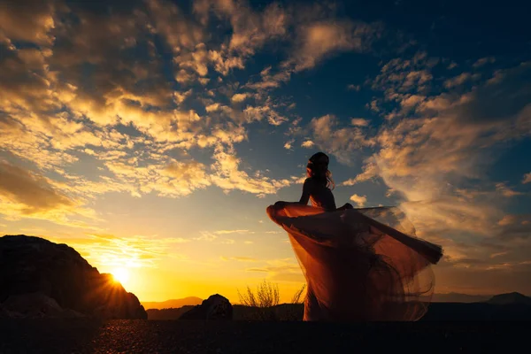 Bruid in een uitwaaierende jurk staat op een berg tegen de achtergrond van een zonsondergang hemel — Stockfoto