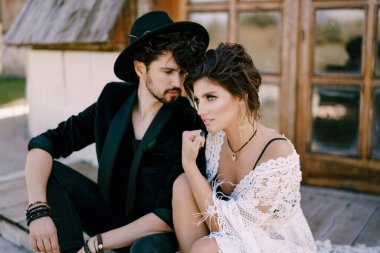 Siyah şapkalı damat, ahşap bir evin kapısında hasır bir elbiseyle gelinin yanında oturuyor.