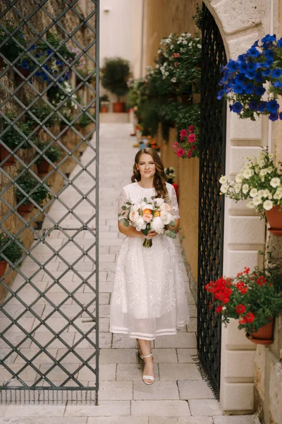 Noiva com um buquê de flores sai de um antigo pátio com um portão decorativo — Fotografia de Stock