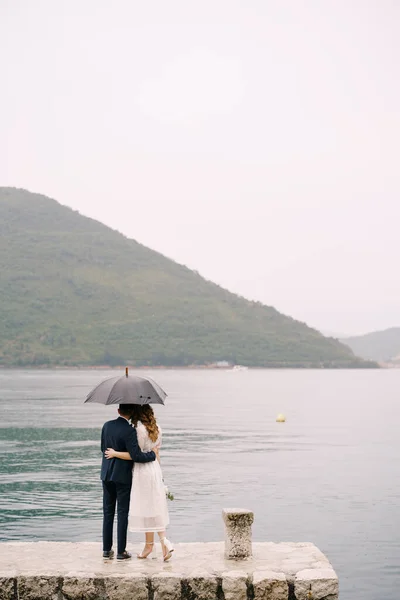 Noiva e noivo estão no cais sob um guarda-chuva. Visão traseira — Fotografia de Stock