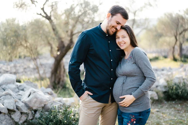 Mujer embarazada sonriente abrazando a un hombre sobre un fondo de árboles —  Fotos de Stock