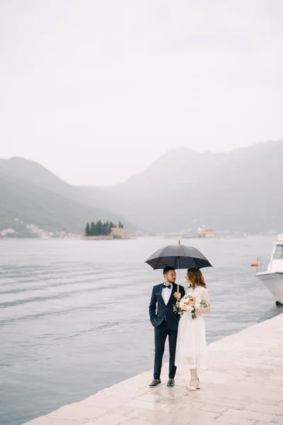Жених и невеста с зонтиком стоят на пирсе у моря на фоне гор — стоковое фото