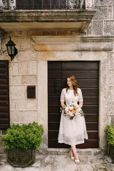 Bride with a bouquet stands at the door of an old stone house — Stock Photo, Image