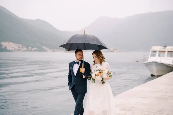 A vőlegény és a menyasszony esernyő alatt sétálnak a mólón. — Stock Fotó