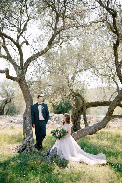 Η νύφη κάθεται σε ένα κορμό δέντρου με ένα μπουκέτο λουλούδια δίπλα στον όρθιο γαμπρό — Φωτογραφία Αρχείου