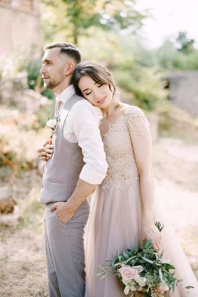 Braut mit Strauß in der Hand umarmt Bräutigam von hinten mit dem Kopf auf seiner Schulter — Stockfoto
