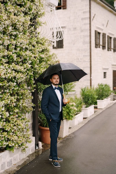Наречений стоїть під парасолькою біля будівлі, переплетеної квітучою зеленню — стокове фото