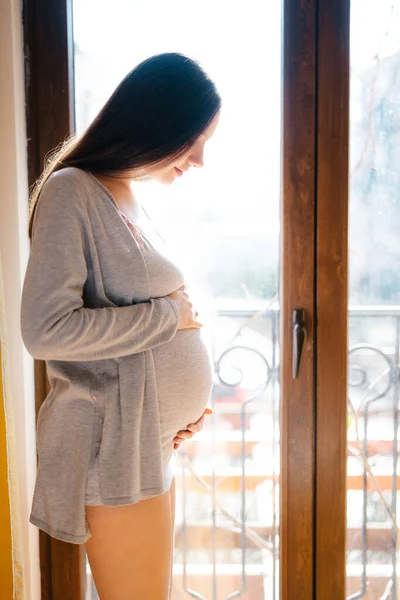 Donna incinta si trova vicino alla porta del balcone accarezzando la pancia — Foto Stock