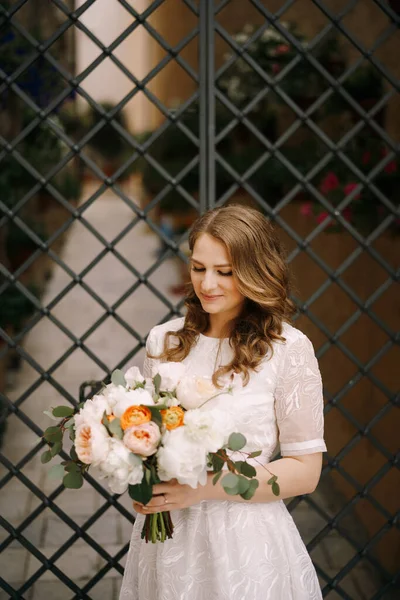 Наречена в білій сукні з букетом квітів стоїть біля декоративних воріт — стокове фото