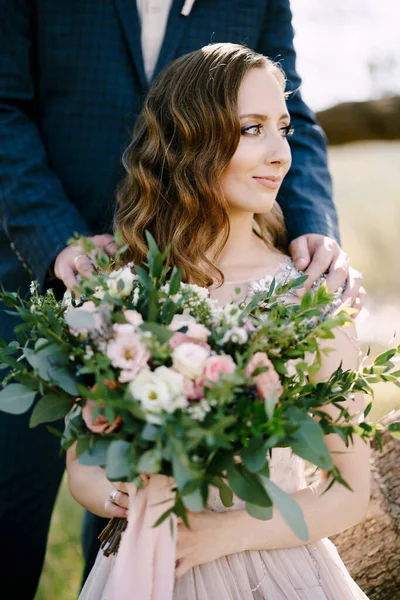 Ο γαμπρός έβαλε τα χέρια του στους ώμους της νύφης καθισμένος σε ένα κορμό δέντρου με ένα μπουκέτο λουλούδια — Φωτογραφία Αρχείου