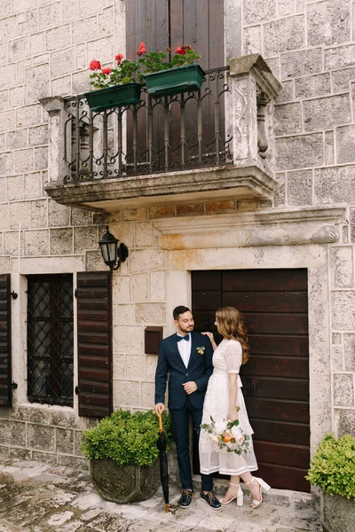 Groom and bride stand near the door of an old stone house with a balcony and shutters — Stock Photo, Image