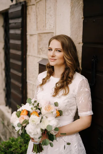 Невеста с букетом опирается на стену у двери старого каменного дома — стоковое фото