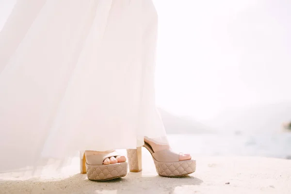 Bruid benen in een witte jurk en beige hoge hakken op de pier. Close-up — Stockfoto