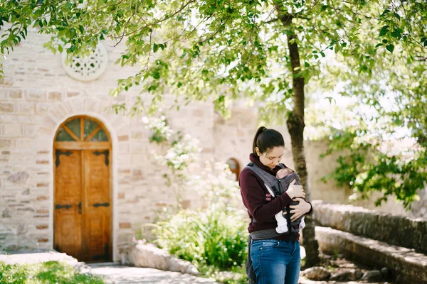 石造りの建物の庭の木の近くに立っている間、母はスリングで赤ちゃんを抱きます — ストック写真