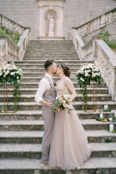 A vőlegény megcsókolja a menyasszonyt, miközben a kőlépcsőn áll a régi villa közelében. — Stock Fotó