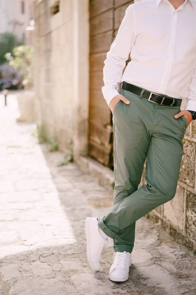 Man in green trousers and a white shirt is leaning against the wall of a stone house. Close-up — Stock Photo, Image