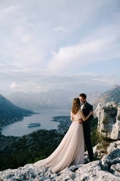 Γαμπρός αγκαλιάζει νύφη από τη μέση σε ένα βραχώδες βουνό με θέα στον κόλπο — Φωτογραφία Αρχείου