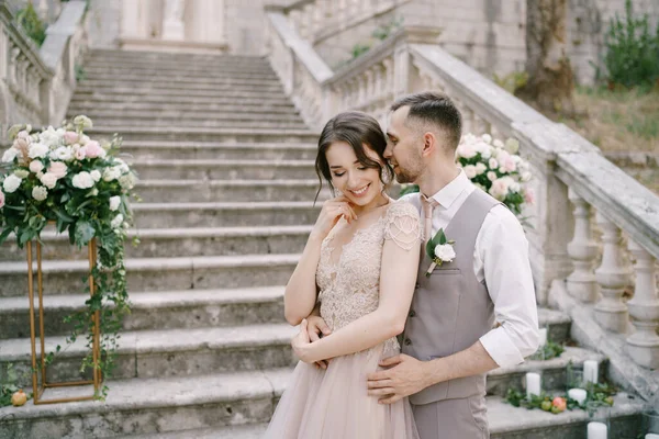 Bräutigam flüstert der Braut ins Ohr, während er sie auf den Steinstufen einer antiken Villa umarmt — Stockfoto
