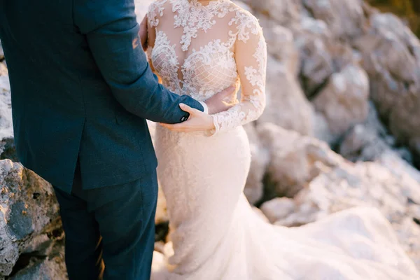 O noivo abraça a noiva pela cintura enquanto está de pé sobre as pedras. Close-up — Fotografia de Stock