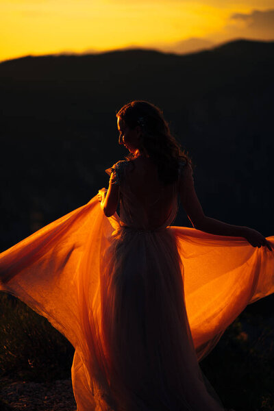 Bride stands holding the hem of the dress on the mountain against the backdrop of the sunset. High quality photo