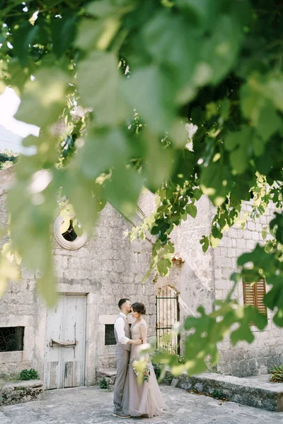 Vista a través del follaje de un árbol en la novia y el novio abrazándose cerca del edificio de piedra —  Fotos de Stock