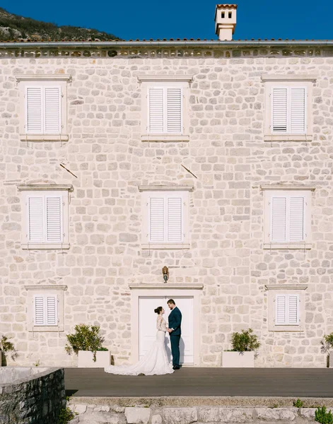Nevěsta a ženich stojí v blízkosti kamenného domu s bílými žaluziemi na oknech — Stock fotografie