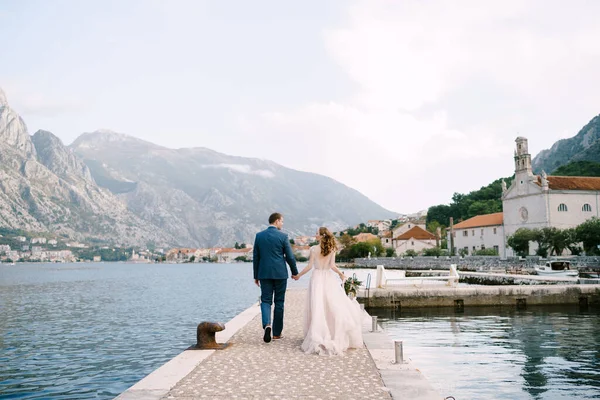 Braut und Bräutigam gehen an der Seebrücke entlang, Händchen haltend vor der Kulisse der Berge. Rückseite — Stockfoto