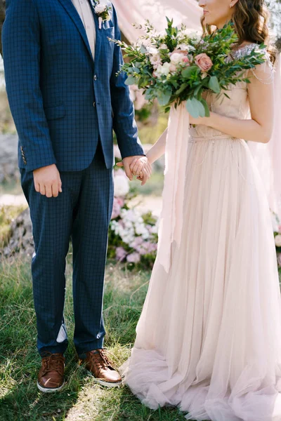 Νύφη με μακρύ φόρεμα με μπουκέτο στέκεται στο χέρι με το γαμπρό σε μπλε καρό κοστούμι — Φωτογραφία Αρχείου