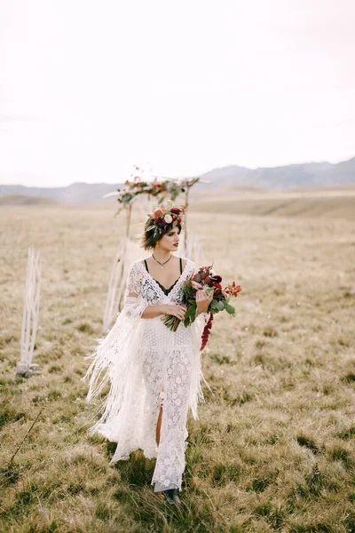 Noiva em uma grinalda com um buquê de flores caminha pelo campo — Fotografia de Stock