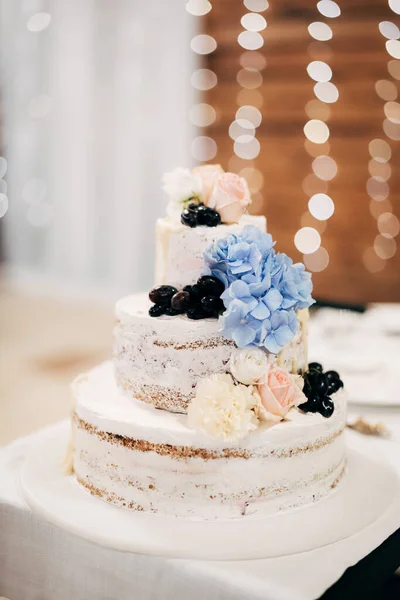 Třístupňový dort zdobený květinami a bobulemi stojí na bílém stojanu na stole — Stock fotografie