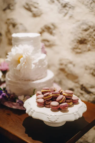 Makarónové dorty leží na stojanu vedle svatebního dortu na stole u kamenné zdi — Stock fotografie