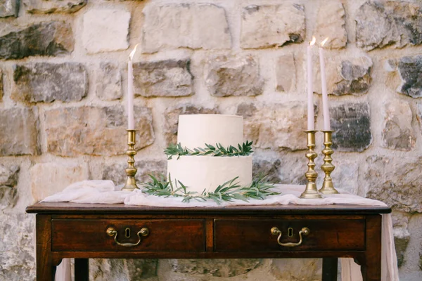 Bunk wedding cake decorated with green branches stands on a wooden table near a stone wall — 图库照片
