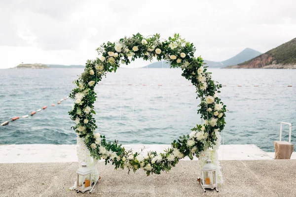 Arco de boda redonda tejida de rosas y hojas verdes se encuentra en el muelle — Foto de Stock