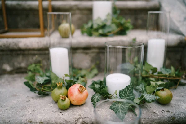 Bílé svíčky v brýlích stojí na kamenných schodech obklopených granátovými jablky a zeleným břečťanem — Stock fotografie