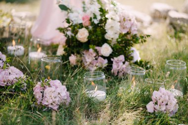 Çiçeklerle süslenmiş cam kavanozlarda yanan mumlar yeşil çimlerde duruyor.