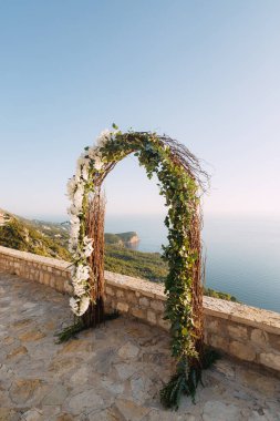 Beyaz orkideler ve dallarla sarmalanmış düğün kemeri denizin üstündeki gözlem güvertesinde duruyor.