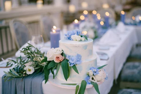 Kue pengantin putih dihiasi dengan bunga dan daun berdiri di atas meja dengan lilin — Stok Foto