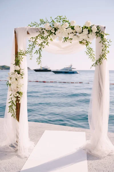 Весільна арка, прикрашена білим тюлем, трояндами і зеленими гілками, стоїть на пірсі біля моря — стокове фото