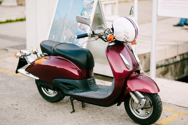 Beyaz kasklı Burgundy scooter sokakta duruyor.