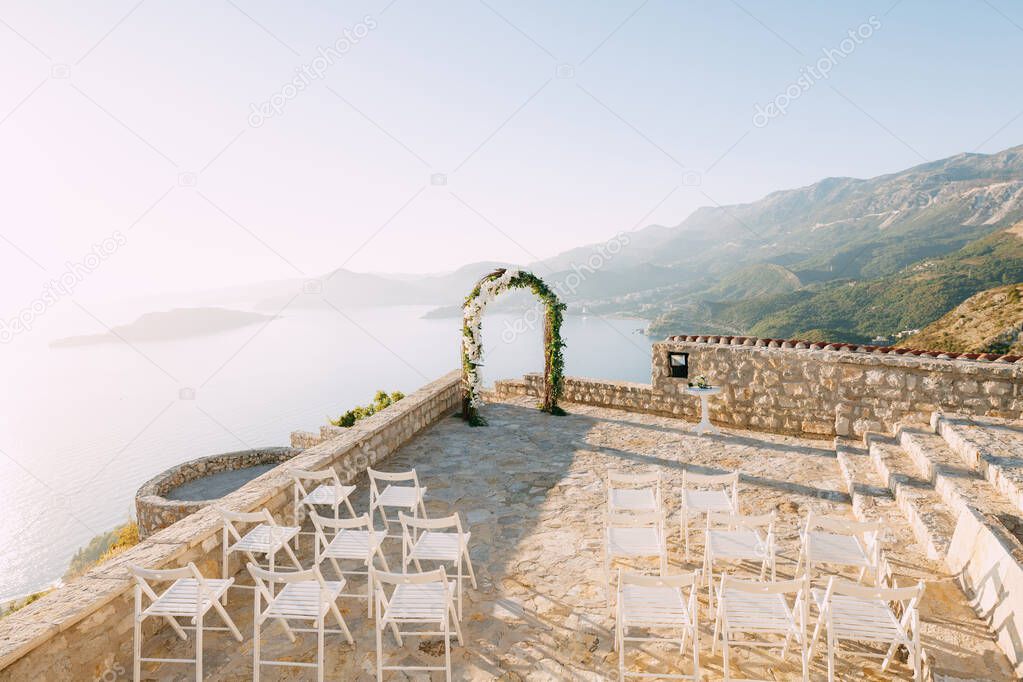 Wedding arch stands on an observation deck above the sea in front of rows of white chairs