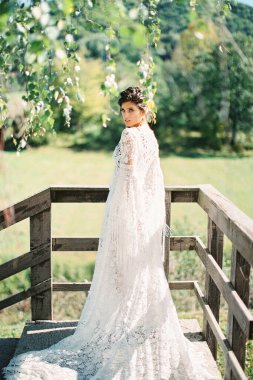 Uzun beyaz hasır elbiseli gelin korkulukların yanındaki terasta duruyor.