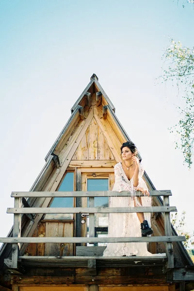 Mariée en robe blanche se tient sur le balcon d'une maison triangulaire appuyée contre une rampe en bois — Photo