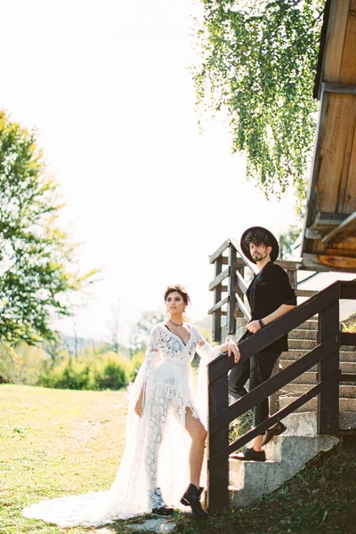 Noiva e noivo estão de pé nos degraus perto dos grades de madeira da casa — Fotografia de Stock