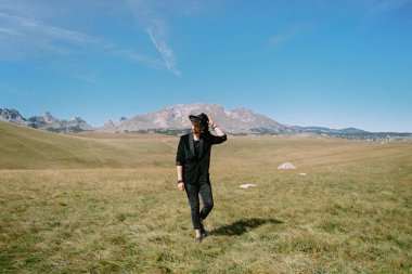 Genç adam elinde şapkasıyla bir vadide yürüyor.