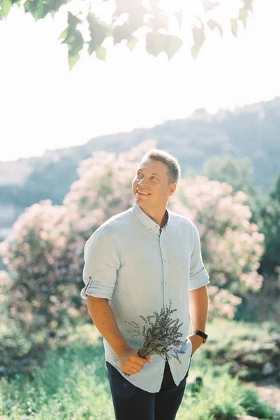Усміхнений чоловік з букетом лаванди в руці стоїть біля квітучого дерева — стокове фото