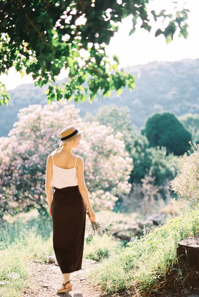 Žena v slaměném klobouku s kyticí levandule kráčí po stezce v horách. Zadní pohled — Stock fotografie